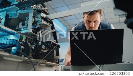 Male Technician in Uniform Setting and Servicing Automated Teller Machine Machine Using Laptop 126203203