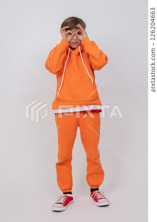 Boy in orange sweatshirt and orange pants making glasses sign with hands. Boy standing on grey background Boy in orange sweatshirt and orange pants making glasses sign with hands. Boy standing on grey background 126204663