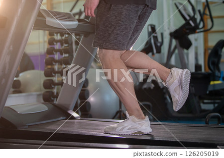 A man is running on a treadmill in a gym. The man is wearing shorts and a white shirt 126205019