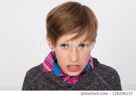 A boy in a striped shirt and a blue scarf looks at the camera with a frowning expression on his face. 126206237