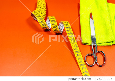 A measuring tape and sewing scissors with a yellow cloth on an orange table. A measuring tape and sewing scissors with a yellow cloth on an orange table. 126209997