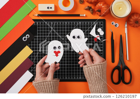 Paper crafts for halloween ghosts. Hands holding finished crafts against a background of diy crafting supplies. Cutting mat, scissors, cutter, tweezers, pencil, eraser, tape, paper. Paper crafts for halloween ghosts. Hands holding finished crafts against a background of diy crafting supplies. Cutting mat, scissors, cutter, tweezers, pencil, eraser, tape, paper. 126210197