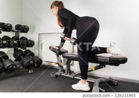 Woman doing dumbbell row on bench in modern gym. Woman doing dumbbell row on bench in modern gym. 126211903