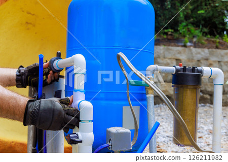 Plumber connects pipes to artesian water well filtration tank during an installation process in backyard. Plumber connects pipes to artesian water well filtration tank during an installation process in backyard. 126211982