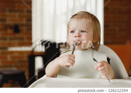 Infant girl or toddler sitting on high chair. Eating food by herself using spoon 126212204