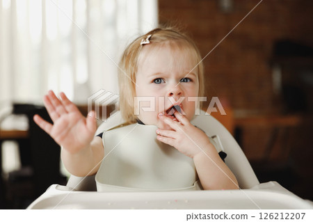 Infant girl or toddler sitting on high chair. Eating food by herself using spoon Infant girl or toddler sitting on high chair. Eating food by herself using spoon 126212207