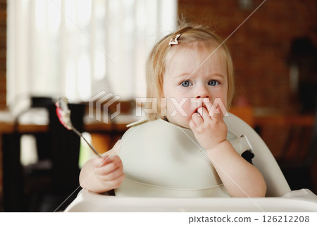 Infant girl or toddler sitting on high chair. Eating food by herself using spoon Infant girl or toddler sitting on high chair. Eating food by herself using spoon 126212208