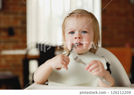 Infant girl or toddler sitting on high chair. Eating food by herself using spoon Infant girl or toddler sitting on high chair. Eating food by herself using spoon 126212209