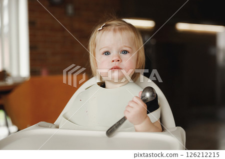 Infant girl or toddler sitting on high chair. Eating food by herself using spoon 126212215