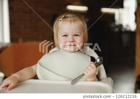 Infant girl or toddler sitting on high chair. Eating food by herself using spoon Infant girl or toddler sitting on high chair. Eating food by herself using spoon 126212216