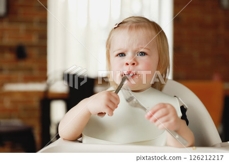 Infant girl or toddler sitting on high chair. Eating food by herself using spoon Infant girl or toddler sitting on high chair. Eating food by herself using spoon 126212217