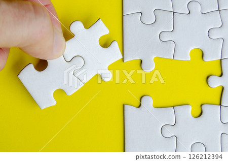 Hand matching white puzzle pieces lying on a vibrant yellow surface highlighting contrast and precise alignment. Hand matching white puzzle pieces lying on a vibrant yellow surface highlighting contrast and precise alignment. 126212394