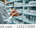 Pharmacist examines blister pack of pills in pharmacy. Shelves are stacked with various medicines. Close-up 126213089