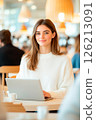Smiling woman sits at cafe table using laptop, embodying relaxed yet focused work environment 126213091