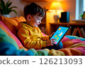 A young child interacts with an educational app on a tablet while cozying up on a couch 126213093