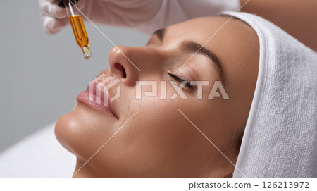 Closeup of glowing skin treatment with dropper applying serum on relaxed woman face wrapped in white towel, showing smooth skin texture 126213972