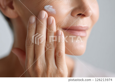 Closeup of woman gently applying antiaging cream on her cheek, showing smooth skin and calm smile with natural light 126213974