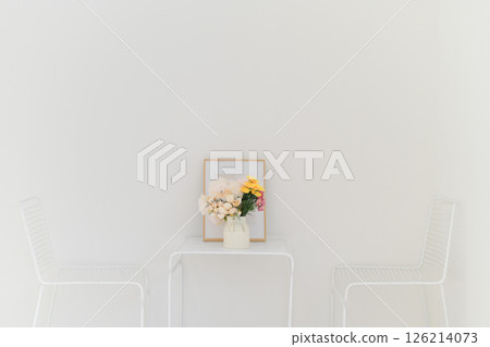 Minimalist white room featuring small table with floral arrangement, framed picture, and two white chairs creating clean and serene atmosphere 126214073
