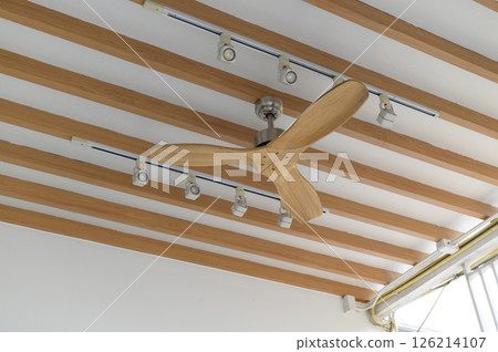 Close up view of modern ceiling fan with wooden blades mounted on ceiling with wooden beams and track lighting, creating warm and stylish atmosphere Close up view of modern ceiling fan with wooden blades mounted on ceiling with wooden beams and track lighting, creating warm and stylish atmosphere 126214107