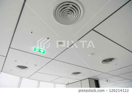 Clean modern office ceiling featuring air vents, recessed lighting, and grid pattern with emergency exit sign glowing green, creating bright and organized atmosphere 126214122