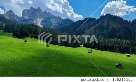 Majestic mountain landscape with lush green fields and rustic cabins under a bright blue sky in the Dolomites 126215193