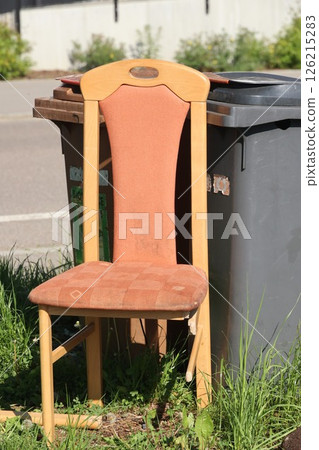 A wooden chair with a brown cushion is sitting on the grass next to a trash can A wooden chair with a brown cushion is sitting on the grass next to a trash can 126215283