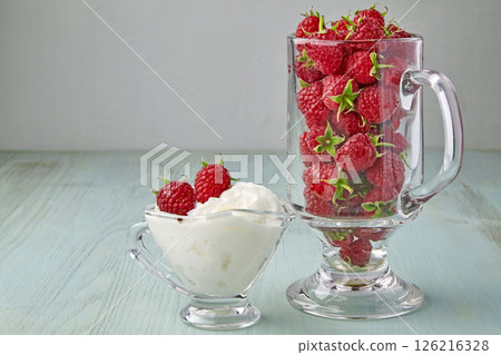 ripe raspberries in a glass goblet and in a sauceboat with cream on a blue wooden table 126216328