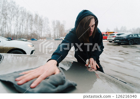 Person cleaning a car hood in a snowy parking lot during a cold winter day Person cleaning a car hood in a snowy parking lot during a cold winter day 126216340