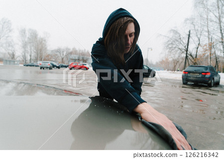 Person cleaning a car hood in a snowy parking lot during a cold winter day 126216342