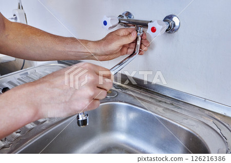 Male hands are tightening locking nut of faucet spout during installation of new dual handle kitchen tap above stainless steel sink with water traces on draining board. 126216386