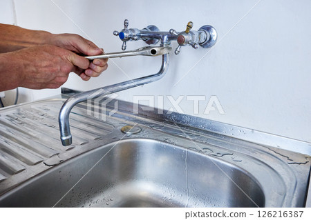 Mounting of faucet spout is performed by plumber using adjustable wrench designed for turning hexagonal fittings during plumbing work, wet stainless steel sink reflects recent activity. Mounting of faucet spout is performed by plumber using adjustable wrench designed for turning hexagonal fittings during plumbing work, wet stainless steel sink reflects recent activity. 126216387