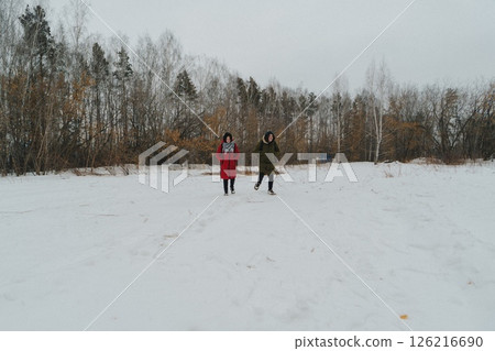 Two individuals stroll across a vast expanse of white snow amidst a nearly bare forest. Two individuals stroll across a vast expanse of white snow amidst a nearly bare forest. 126216690