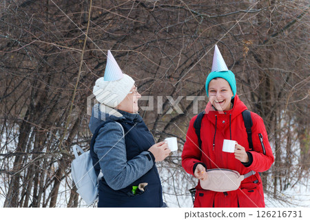 Two friends stand joyfully in a snowy forest, each wearing colorful party hats. Two friends stand joyfully in a snowy forest, each wearing colorful party hats. 126216731