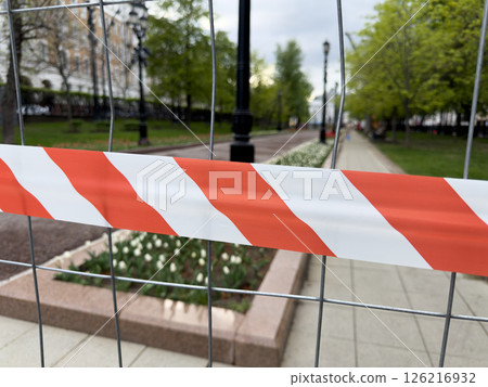 Warning tape on metal fence blocks access to landscaped park area during maintenance, symbolizing safety, restricted entry, and temporary closure for improvement works Warning tape on metal fence blocks access to landscaped park area during maintenance, symbolizing safety, restricted entry, and temporary closure for improvement works 126216932