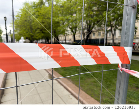 Red and white warning tape stretched across metal fence indicates restricted access in a public park area. Safety measure marks the boundary of an ongoing maintenance zone Red and white warning tape stretched across metal fence indicates restricted access in a public park area. Safety measure marks the boundary of an ongoing maintenance zone 126216933