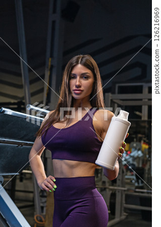 fit young woman poses confidently in a stylish purple crop top and matching leggings while holding a sports bottle. The gym behind her features workout equipment creating an energetic atmosphere. fit young woman poses confidently in a stylish purple crop top and matching leggings while holding a sports bottle. The gym behind her features workout equipment creating an energetic atmosphere. 126216969