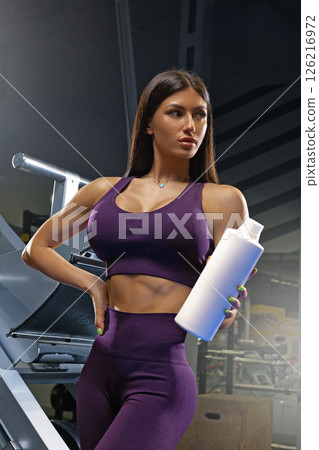 strong woman dressed in a purple workout outfit stands confidently in a gym holding a water bottle. She embodies fitness and strength as she prepares for her exercise routine. 126216972