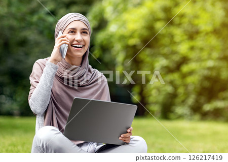 Positive arab girl freelancer working at park, using laptop and talking with clients on phone, copy space Positive arab girl freelancer working at park, using laptop and talking with clients on phone, copy space 126217419