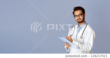 Portrait of young Indian male doctor writing on clipboard over lilac background. Panorama Portrait of young Indian male doctor writing on clipboard over lilac background. Panorama 126217421