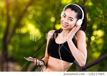 Asian female runner having break in training, listening music with smartphone and headphones outdoors, copy space Asian female runner having break in training, listening music with smartphone and headphones outdoors, copy space 126217469