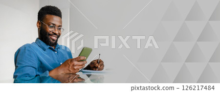 A young African American manager works in a contemporary office space, using a smartphone while looking satisfied. He holds a pen and jotting down notes as he focuses on his tasks, copy space 126217484