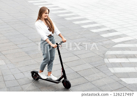 High angle view of cheerful casual girl riding black stand-up scooter on brick road, empty space for ad or text 126217497