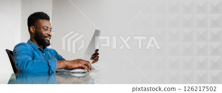 A young African American businessman reviews important documents while seated at a sleek desk. He is focused on his work in a bright, contemporary office environment, copy space A young African American businessman reviews important documents while seated at a sleek desk. He is focused on his work in a bright, contemporary office environment, copy space 126217502