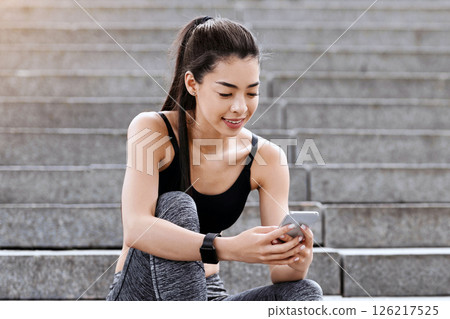 Fit asian girl using smartphone while sitting on stone stairs after training outdoors, resting after fitness workout in city park, free space 126217525
