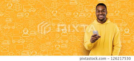 A cheerful buyer stands against a vibrant yellow background filled with delivery and shopping icons. He holds a smartphone, ready to complete an online purchase for a service. A cheerful buyer stands against a vibrant yellow background filled with delivery and shopping icons. He holds a smartphone, ready to complete an online purchase for a service. 126217530