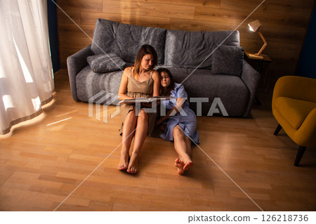 Mother and daughter reading book together sitting on floor in living room. Girl looking bored and tired Mother and daughter reading book together sitting on floor in living room. Girl looking bored and tired 126218736