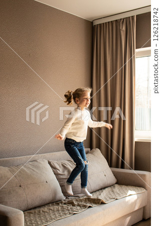 4 years old blonde girl joyfully jumps on couch at home, giggling with excitement. Candid childhood moment of fun and rebellion when parents are not looking. 126218742