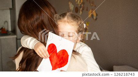 Little girl proudly gives her mom self-made colorful greeting card, sharing a tender embrace. Perfect for themes of parenting, love, and handmade gifts. Little girl proudly gives her mom self-made colorful greeting card, sharing a tender embrace. Perfect for themes of parenting, love, and handmade gifts. 126218743