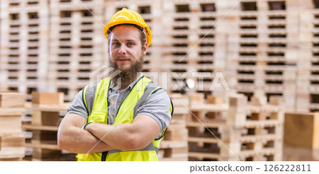 Portrait man worker wearing safety uniform and yellow hard hat standing crossed arms looking at camera smile surrounded piles of pallets background in wooden warehouse. 126222811