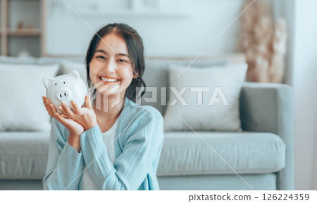 Young Asian woman holding piggy bank and sitting on the floor at home 126224359